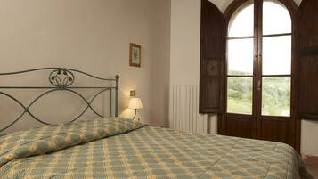 A bed with a checkered bedspread, a bedside lamp, a framed picture on the wall, and a large arched window with a view of greenery.