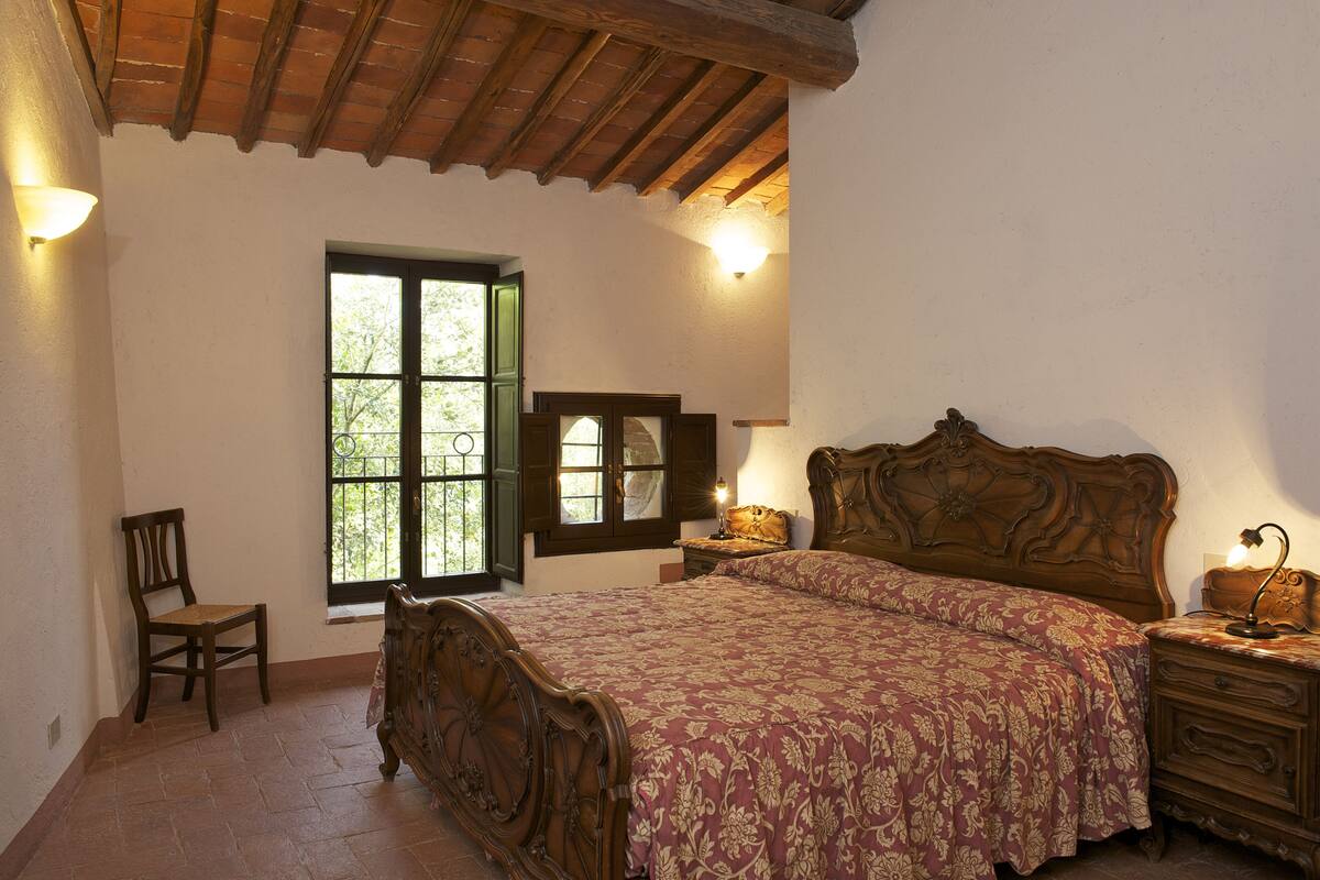 A bedroom with a wooden bed, a chair, a nightstand, and a window with shutters.