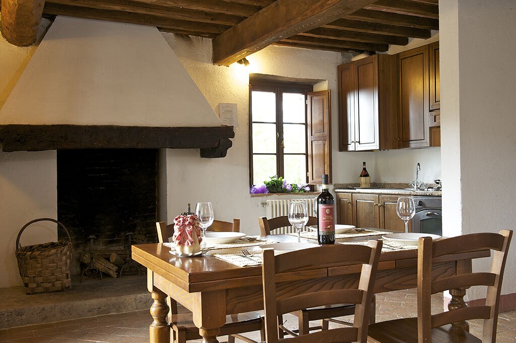 Une salle à manger rustique avec une table en bois dressée pour quatre personnes, une cheminée et une vue sur la cuisine.