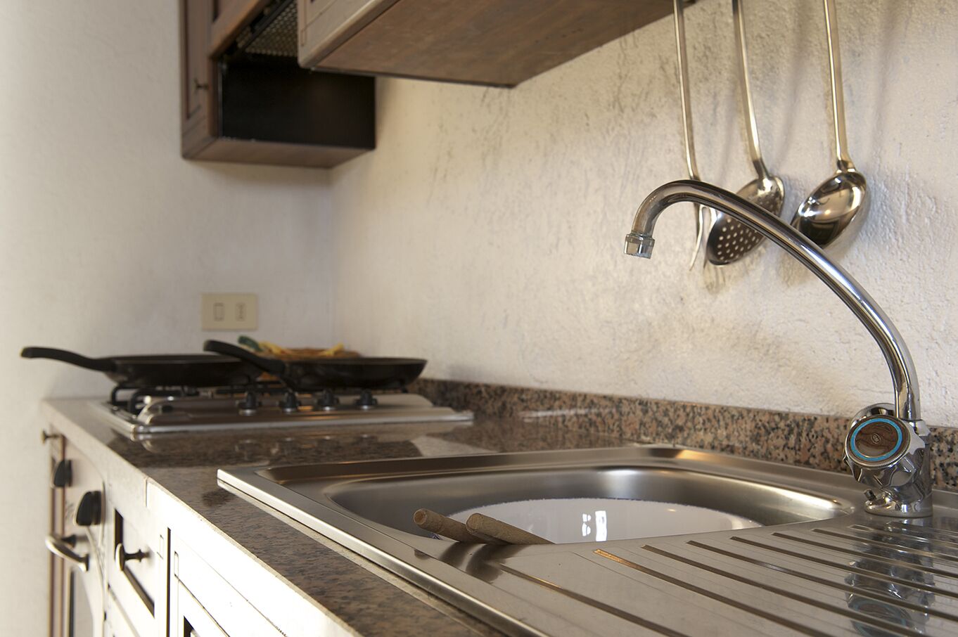 A kitchen with a stainless steel sink, a stove with a frying pan, and hanging utensils.