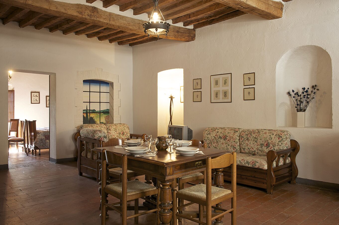 Un comedor con una mesa de madera dispuesta para cuatro personas, rodeada de sillas de madera. Hay un sofá floral contra la pared y un cuadro sobre la mesa.