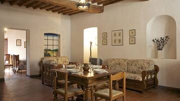 Un comedor con una mesa de madera dispuesta para cuatro personas, rodeada de sillas de madera. Hay un sofá floral contra la pared y un cuadro sobre la mesa.