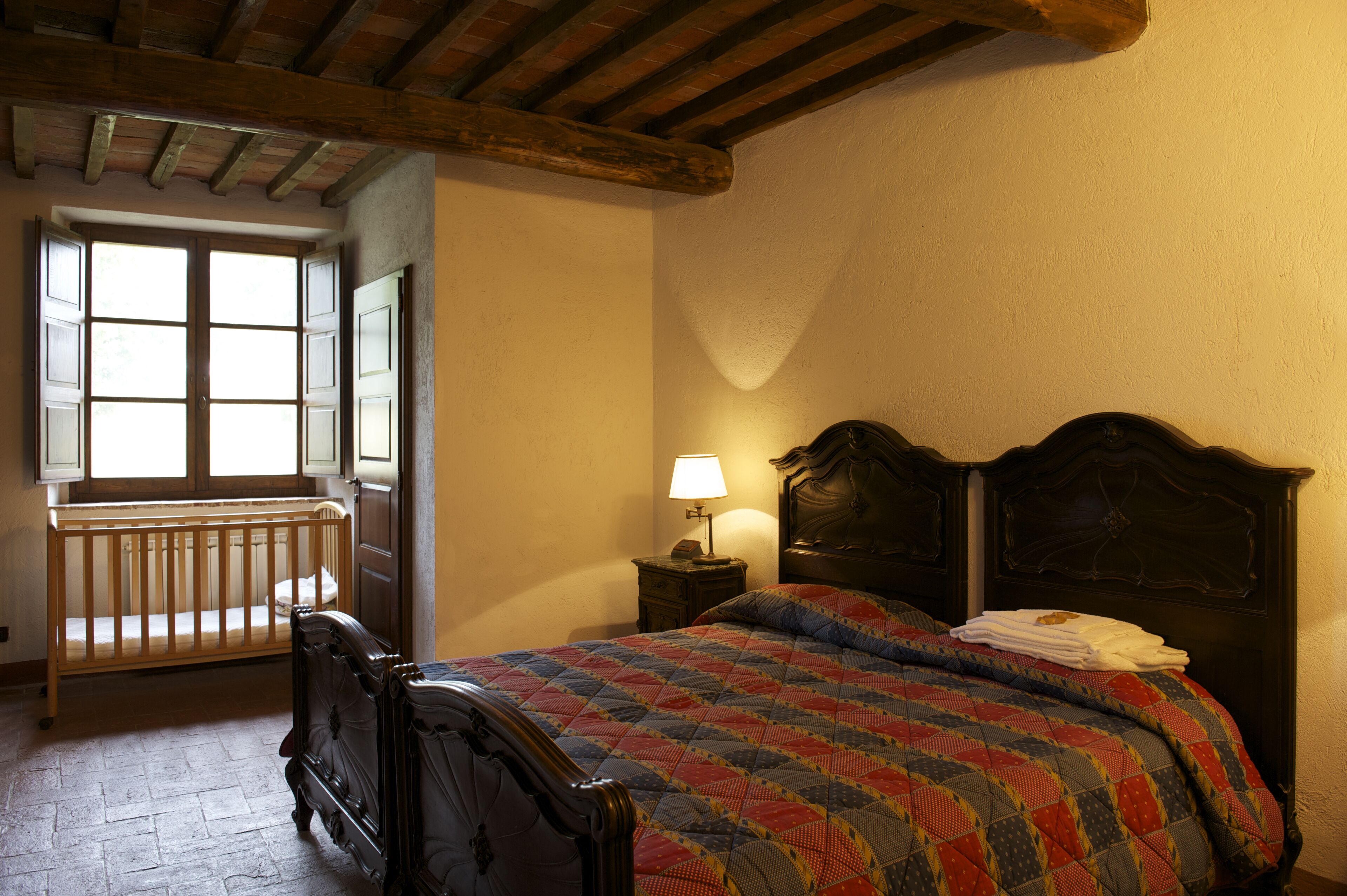 A bedroom with a wooden crib, a bed with a quilt, a nightstand with a lamp, and a window with a view of the outside.