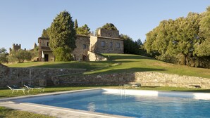 A swimming pool with lounge chairs, a stone wall, and a large building with a dome in the background. - Castello di Modanella (Rapolano Terme)