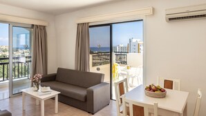 A modern living room with a grey sofa, a small white table, and a dining area with a white table and chairs. A large window offers a view of the city and the sea. - Seagull Hotel Apartments (Paralimni)
