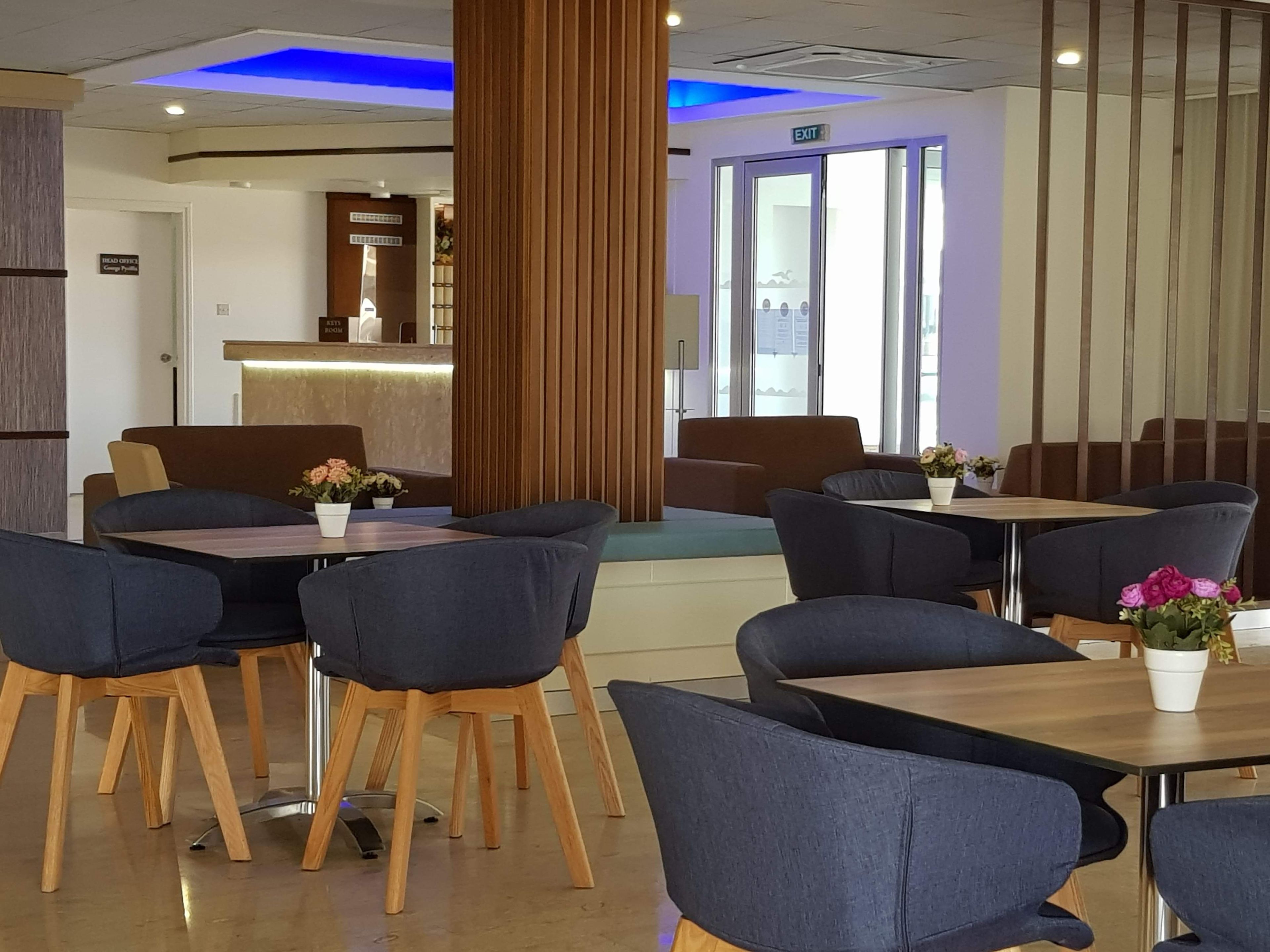 Un hall d’hôtel moderne avec un plancher en bois, des fauteuils bleus et des tables. On y trouve un comptoir de réception avec une cloison en verre et un panneau indiquant la sortie.