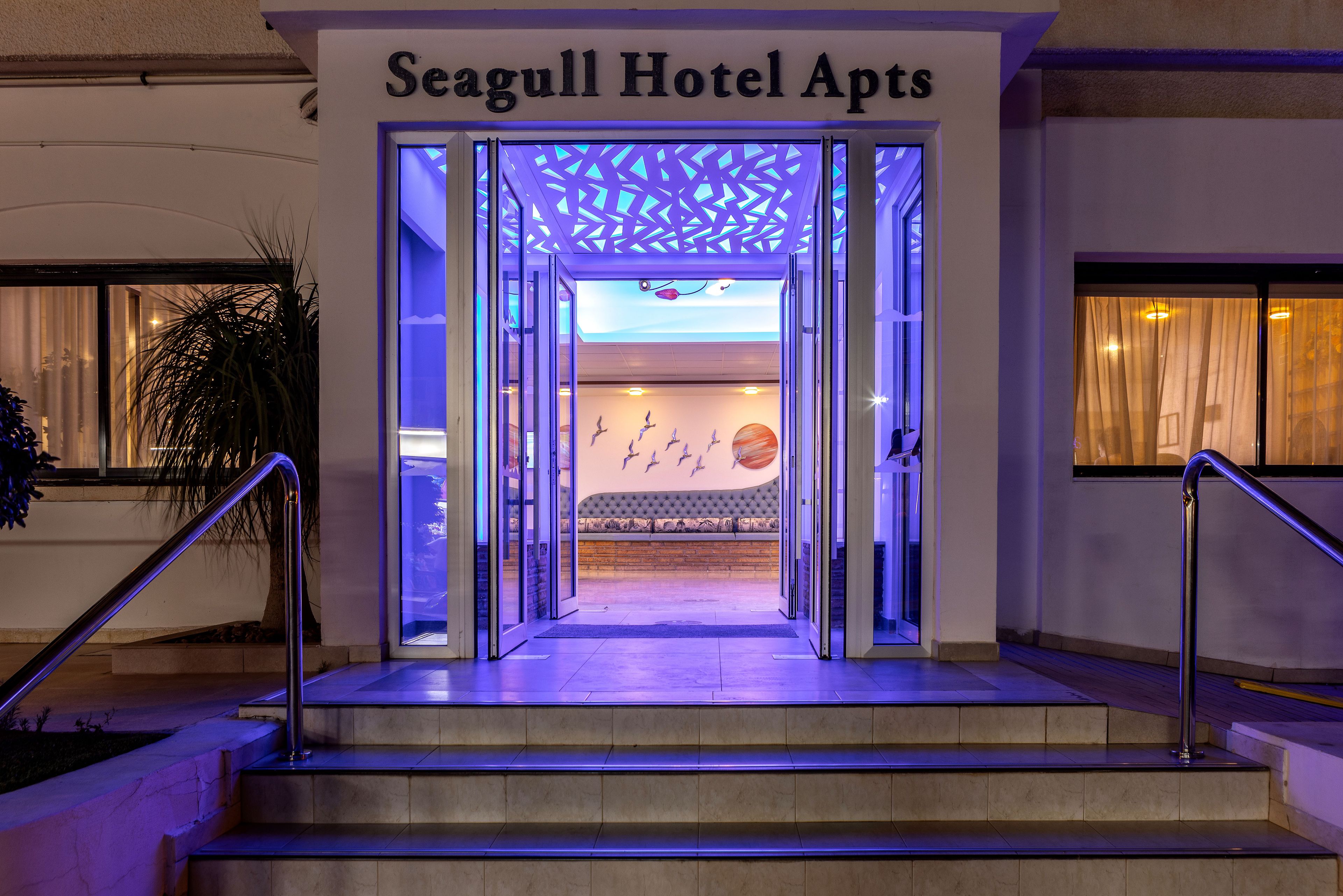 La entrada a Seagull Hotel Apts, con un diseño moderno, presenta una puerta de cristal y una escalera que conduce a la recepción.