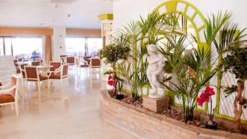 Un vestíbulo amplio con un jardín interior central que incluye una estatua y diversas plantas, rodeado de zonas de estar y grandes ventanales.