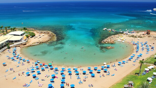 An aerial view of a beach with numerous blue umbrellas and sun loungers, a clear blue sea, and a rocky shoreline. - Seagull Hotel Apartments (Paralimni)