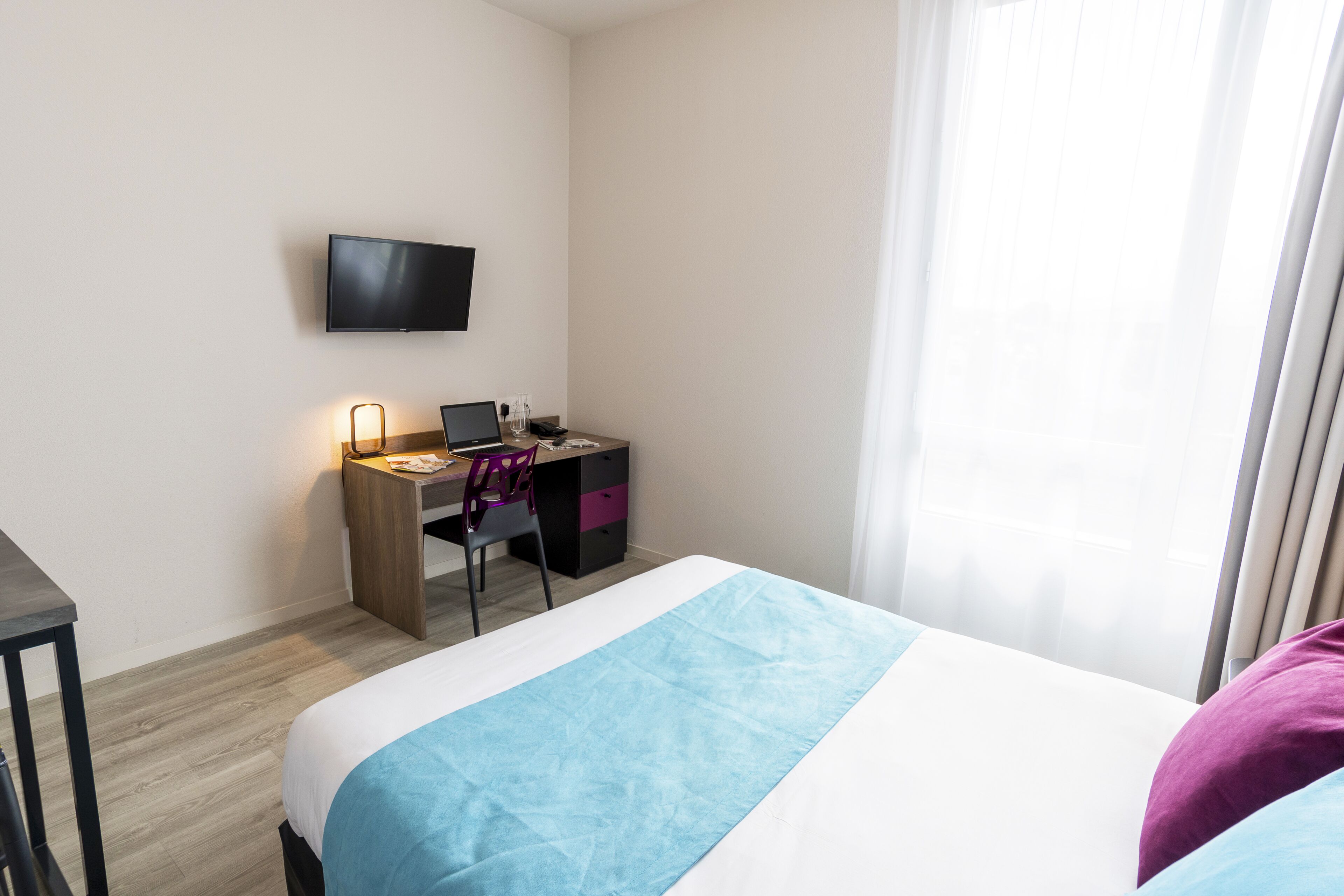 A hotel room with a bed, a desk with a computer, a chair, and a TV on the wall.