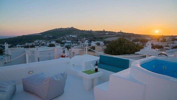 Une terrasse sur le toit, avec des meubles blancs, une piscine bleue et une vue sur la ville et les collines au coucher du soleil.