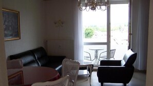 Un salon avec un canapé en cuir noir, des fauteuils en osier blancs, une table ronde et un fauteuil noir. Une fenêtre offre une vue sur la verdure à l’extérieur.