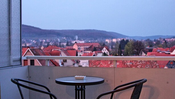 Un balcon avec deux fauteuils en osier et une petite table ronde, d’où l’on peut admirer une ville avec des bâtiments aux toits rouges et des collines à l’horizon.