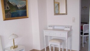 Une coiffeuse blanche avec un miroir et des tiroirs, une petite table de chevet avec une lampe, et un tableau représentant un paysage montagneux.