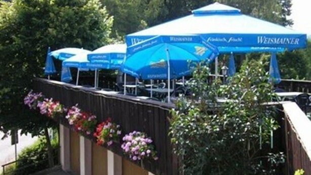 Een rij blauwe parasols met het Weissmainer logo, een houten hek met bloemen en groen op de achtergrond.