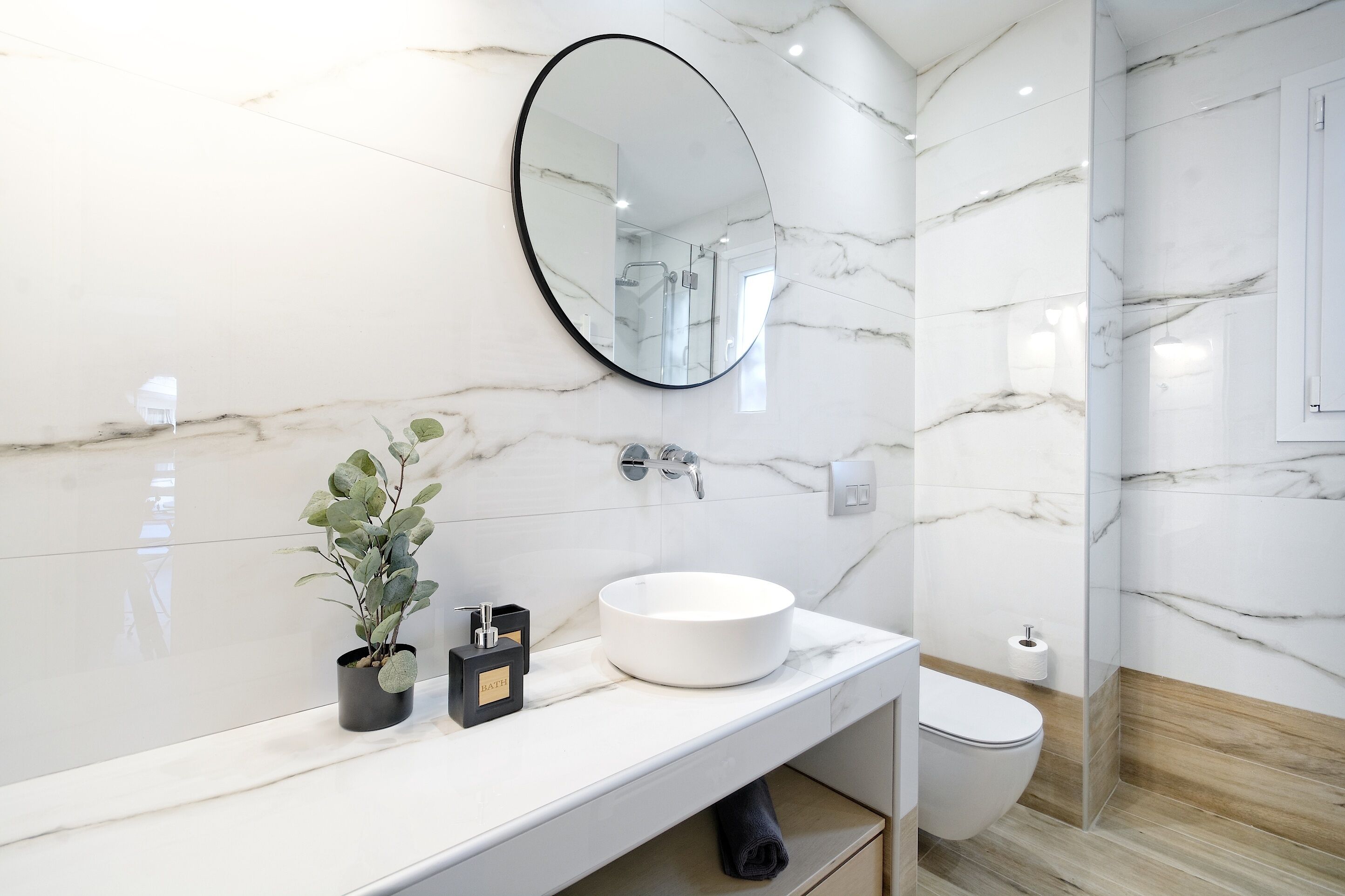 A modern bathroom with a white marble sink, a round mirror, and a potted plant.