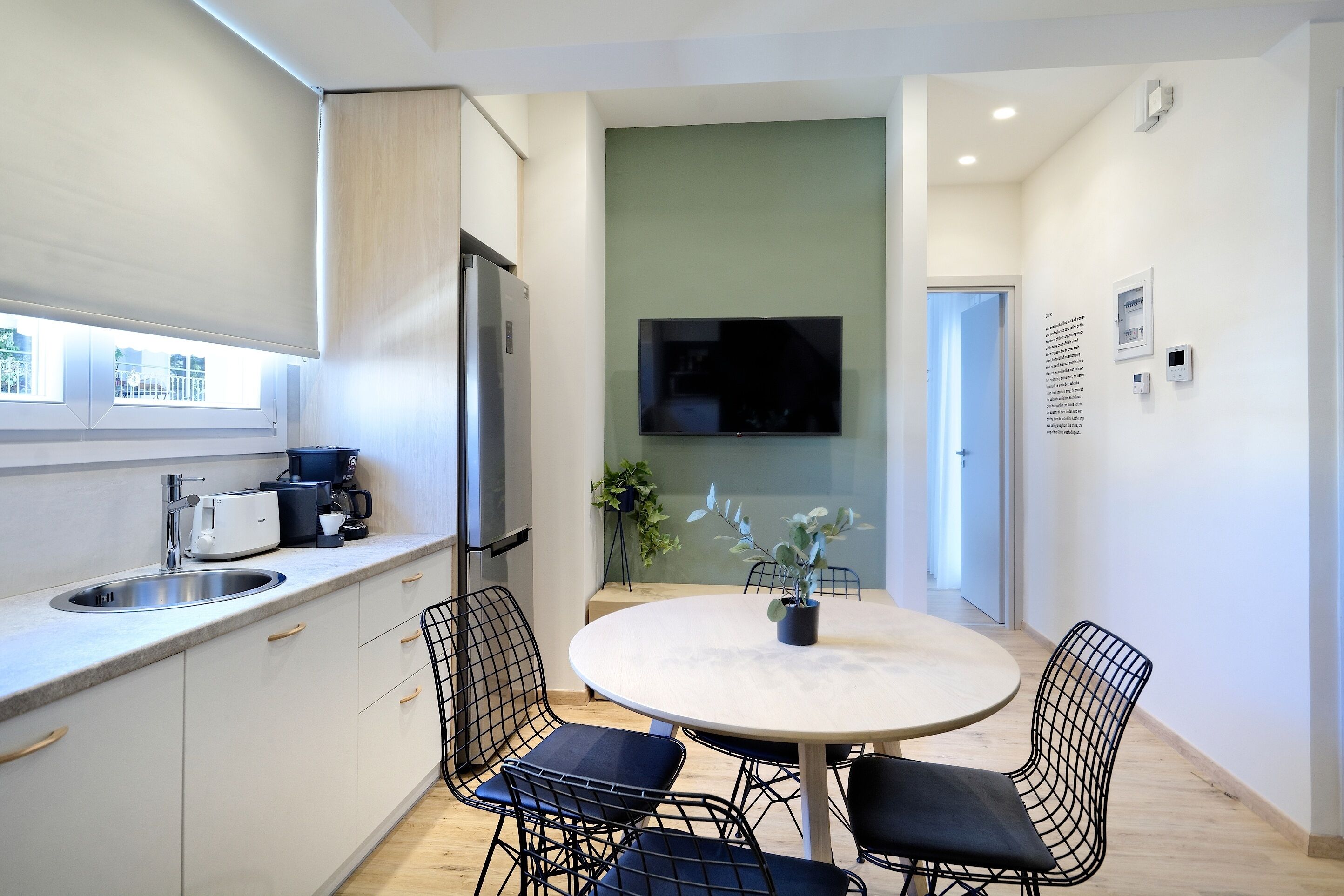 A modern kitchen with a built-in refrigerator, a coffee machine, and a sink. There is a round table with four chairs and a potted plant on it.