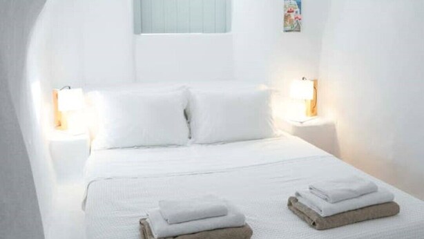 Une chambre à coucher blanche, dotée d’un plafond en arc, comprenant un lit, deux tables de chevet et une fenêtre à cadre bleu.