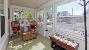 A sunroom with large windows, wicker chairs, a wooden bench, and a decorative chest.