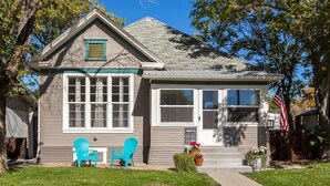 A two-story house with a front porch, two blue Adirondack chairs, and a small garden with potted plants.
