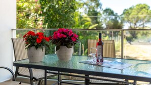 A glass-top table with a bottle of wine, two potted plants with red flowers, and a glass on a balcony with a view of greenery. - Nancy's Luxury Apartments (Zakynthos)