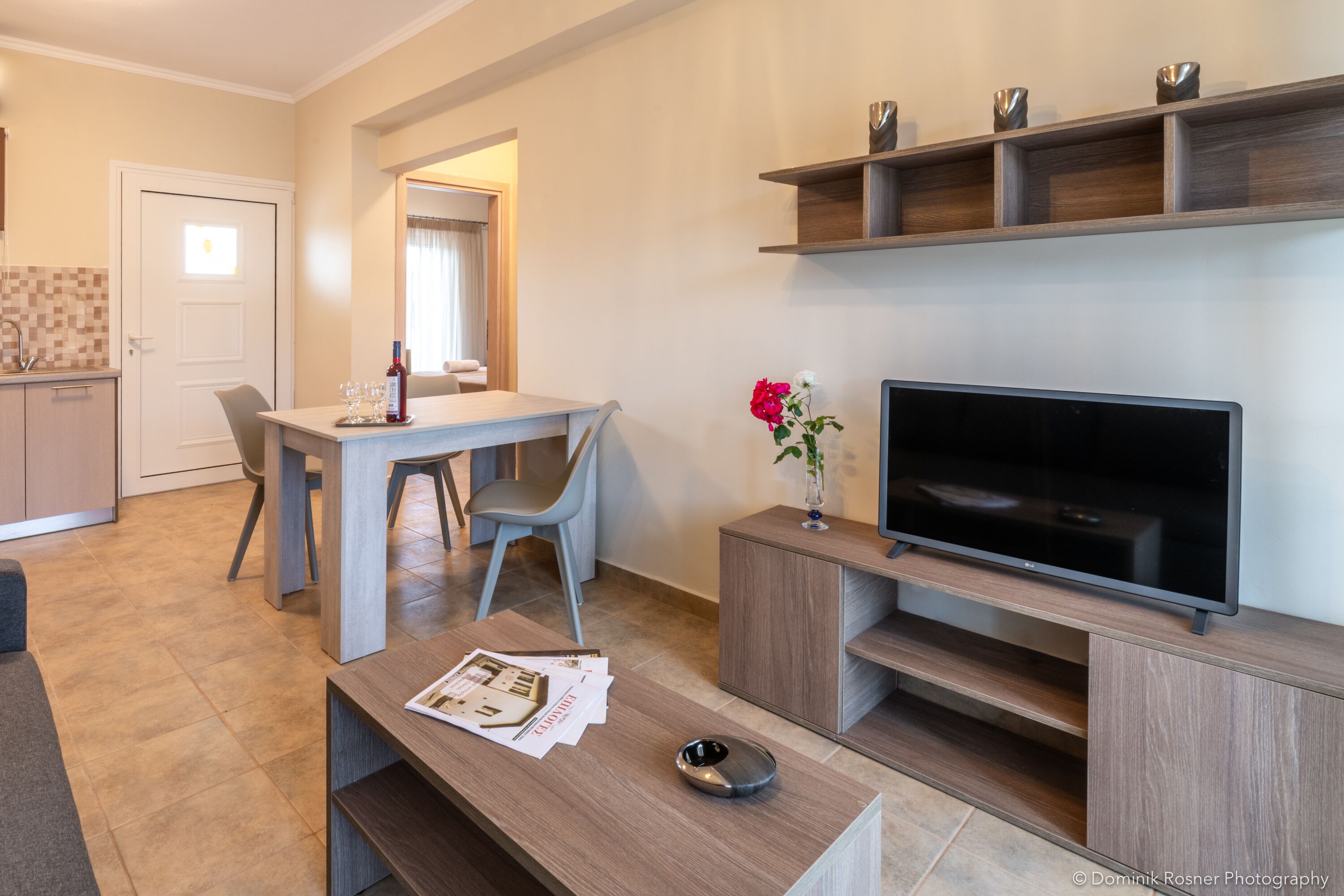 A modern living room with a flat-screen TV, a dining table with chairs, a coffee table with magazines, and a TV stand with decorative items.