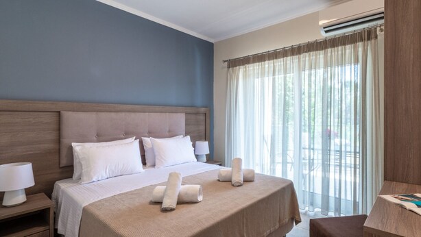 A hotel room with a large bed, two bedside tables with lamps, a wooden headboard, and a view of the outdoors through sheer curtains. - Nancy's Luxury Apartments (Zakynthos)