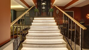 A staircase with a wooden handrail and black balusters, leading to a dark door with a glass panel and two potted plants. - The Fox Hotel (Istanbul)