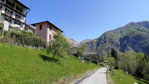 Une route de campagne mène à un bâtiment doté d’un balcon, entouré de collines verdoyantes et de montagnes.