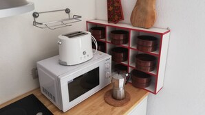 Une cuisine avec un micro-ondes blanc, un grille-pain et une machine à café posés sur le plan de travail. On y trouve des étagères avec des récipients marron et un objet décoratif rouge et noir accroché au mur.