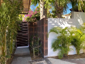 Um caminho leva a um portão com uma escadaria de madeira, cercado por vegetação exuberante e uma parede branca.