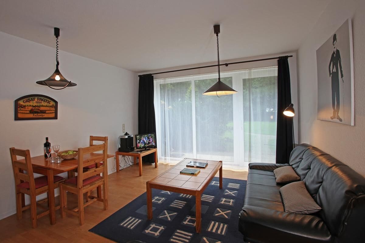 Un salon comprenant une table à manger en bois, des chaises, un canapé et une table basse. On y trouve deux lampes suspendues, un téléviseur et une fenêtre avec des rideaux.