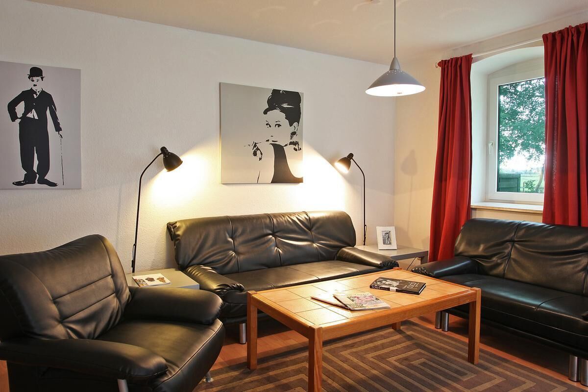 A modern living room with two black leather sofas, a wooden coffee table, and two floor lamps. There are two wall-mounted artworks, one depicting a man and the other a woman. A window with red curtains is visible, and a lamp is on the table.