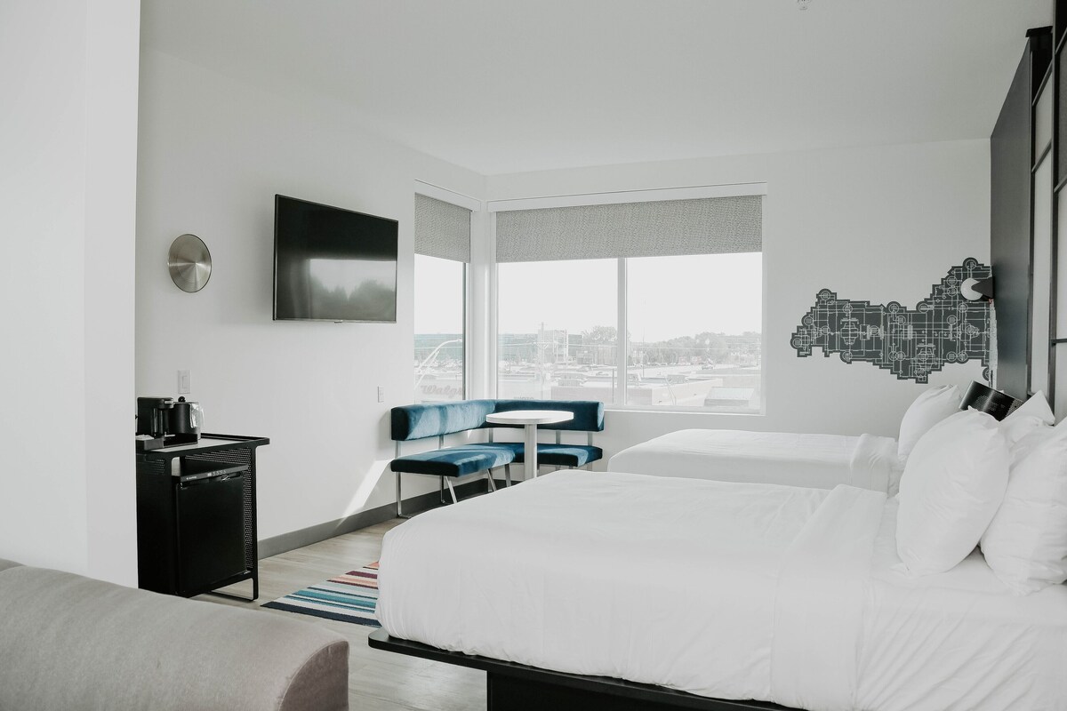 A modern hotel room with a large bed, two seating areas, a flat-screen TV, and a city view.