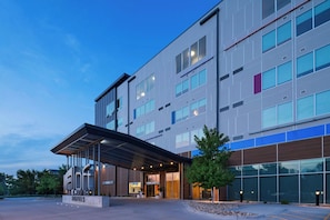 A modern hotel with a covered entrance, large windows, and a landscaped area with trees.