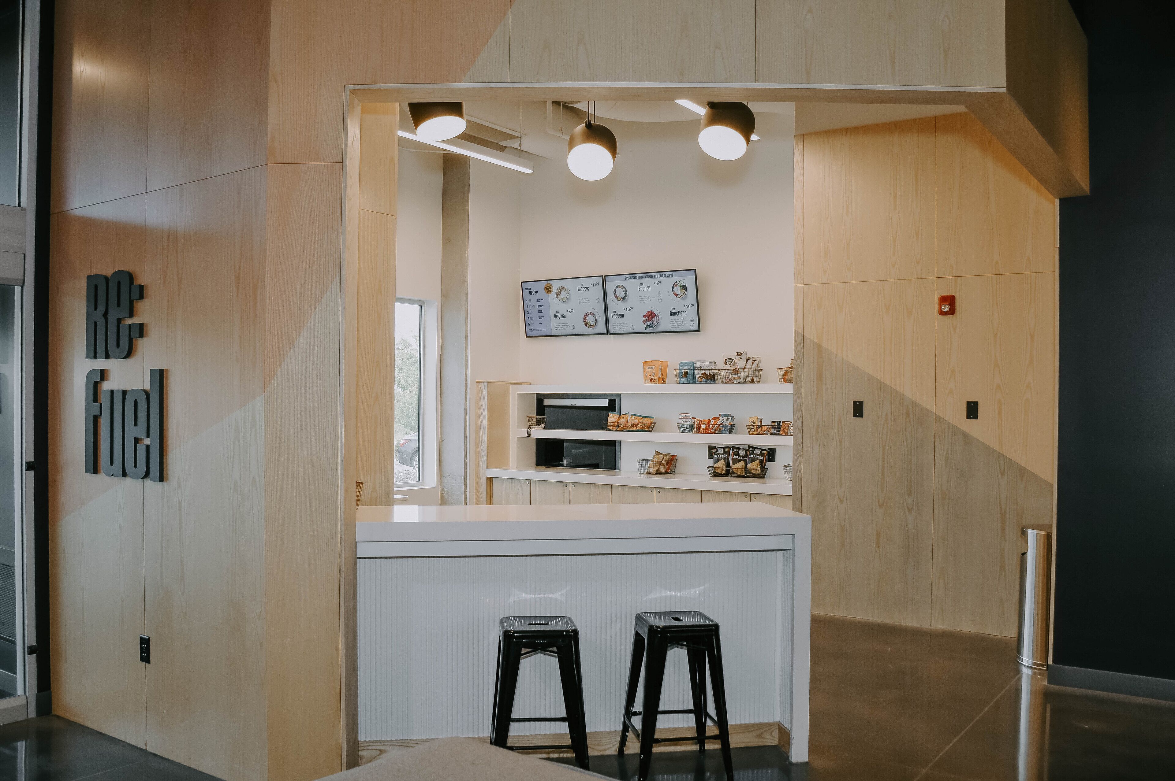 A modern kitchen with wooden cabinetry, a white countertop, and black stools. There are overhead lights and a menu on the wall.