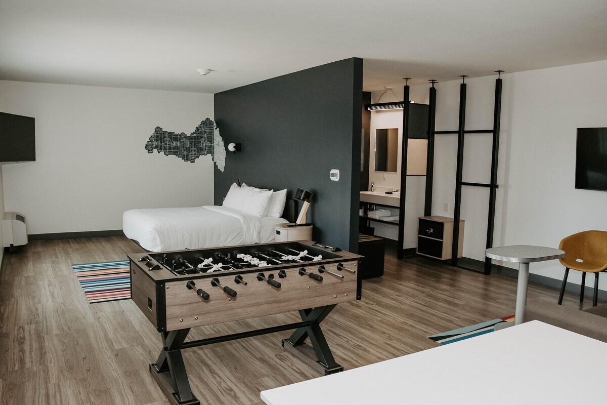 A modern hotel room with a bed, a foosball table, a small table, and a chair.