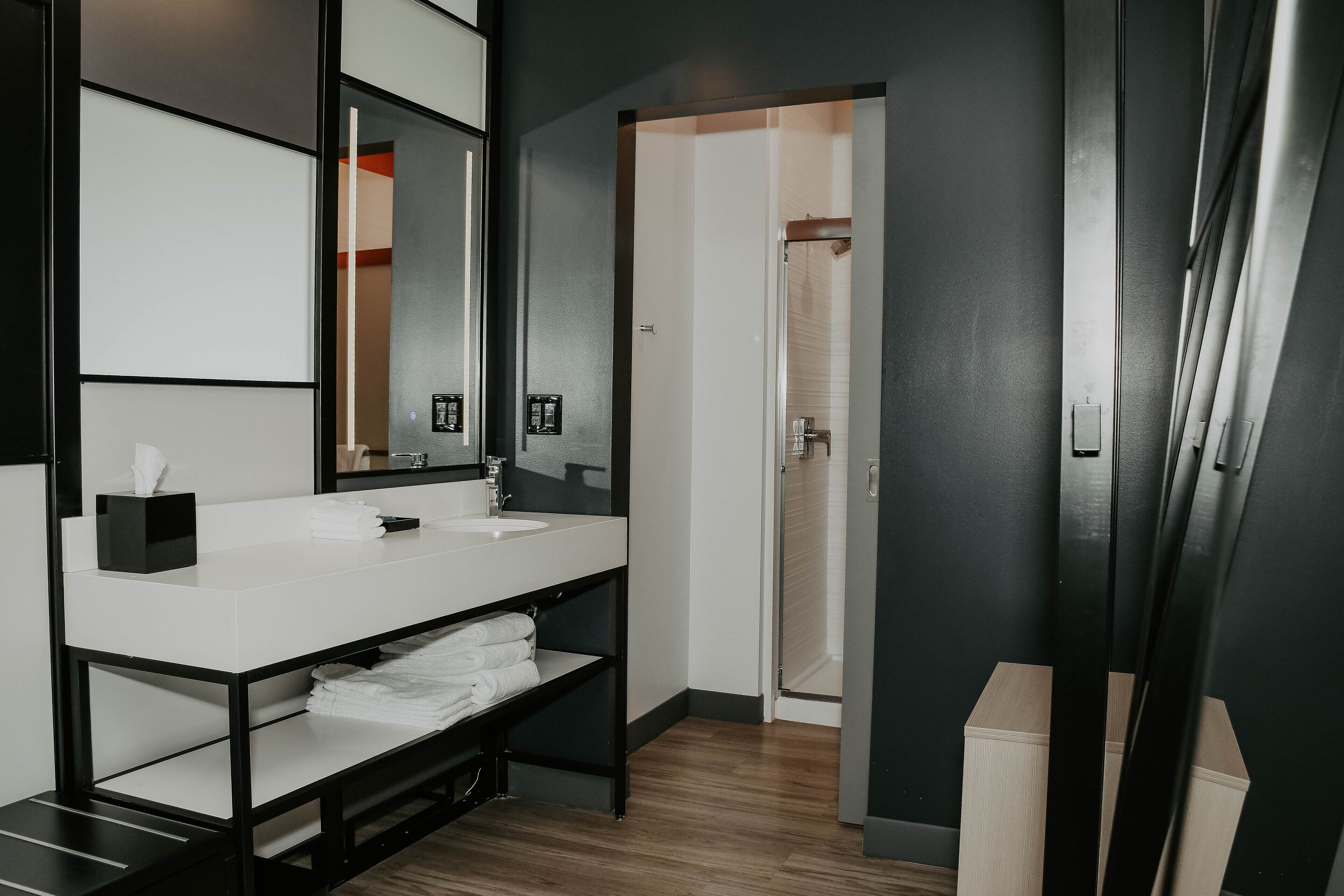 Un baño moderno con un lavamanos blanco, encimera negra y un espejo grande. Se observa un toallero con toallas dobladas y un cubo de basura negro.