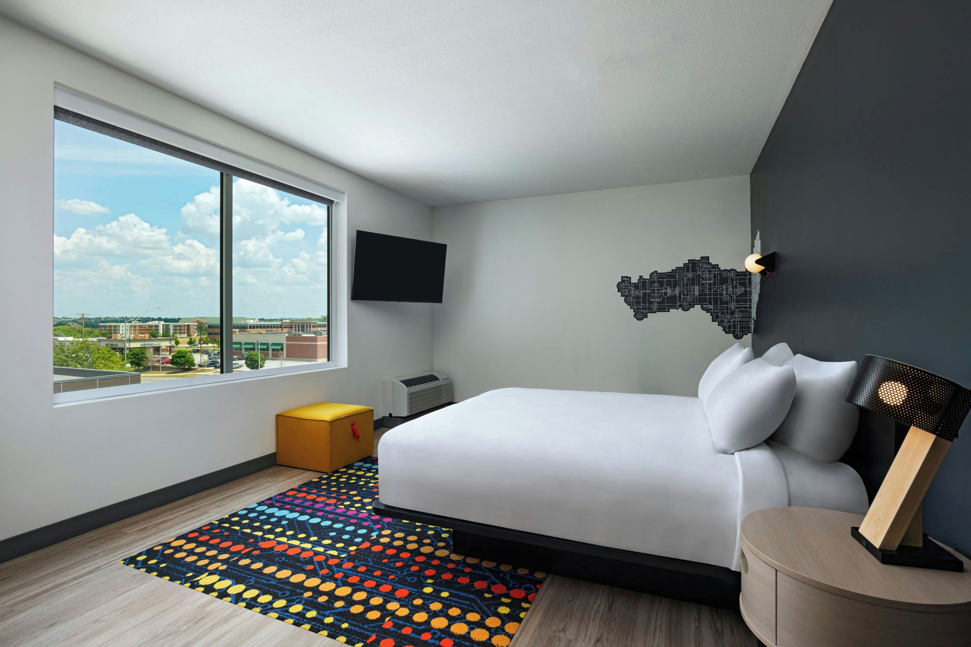A modern hotel room with a large window, a bed, a flat-screen TV, a unique wall art piece, and a colorful rug.
