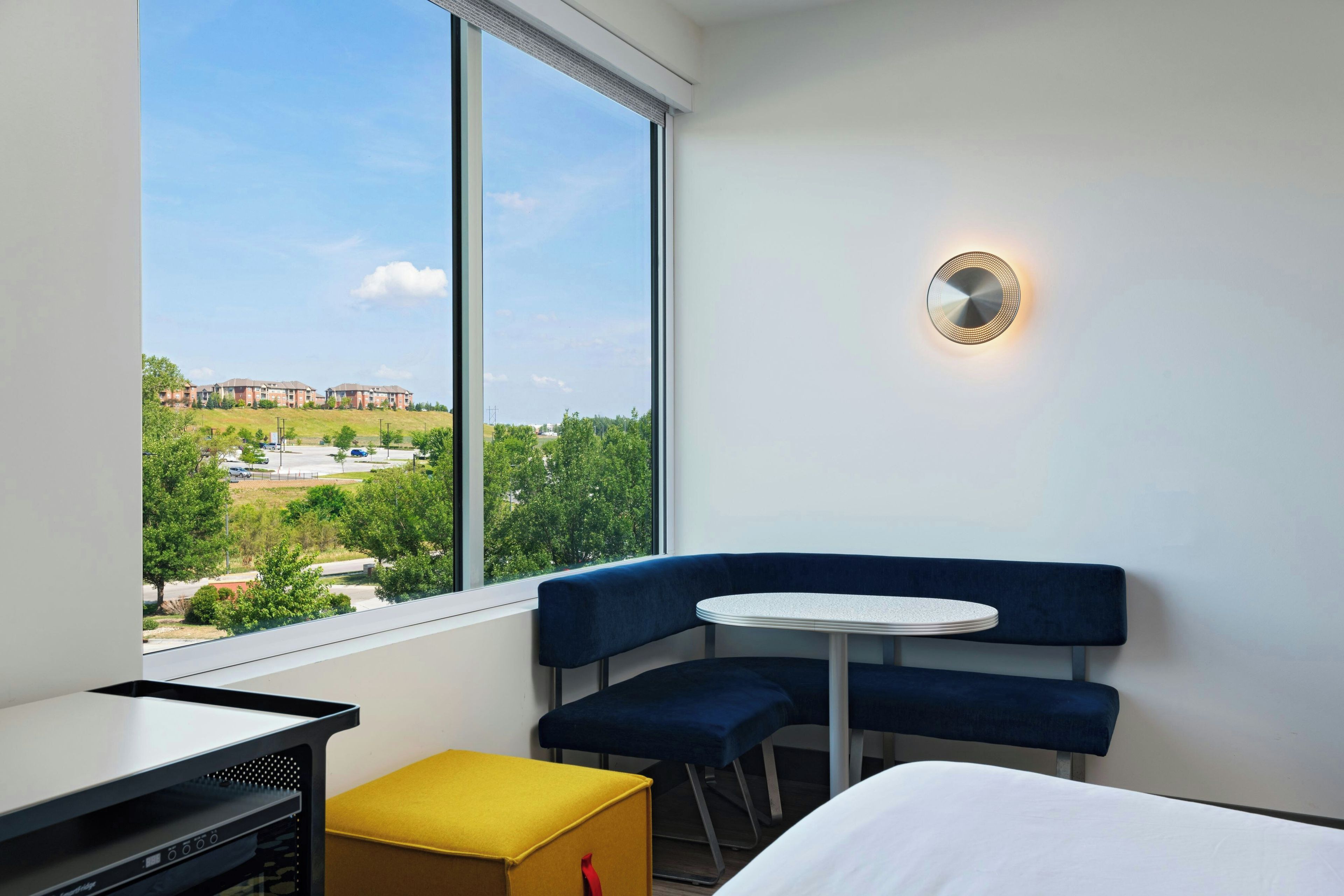 A hotel room with a large window, a round table, a blue sofa, and a small yellow ottoman.