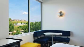 A hotel room with a large window, a round table, a blue sofa, and a small yellow ottoman.