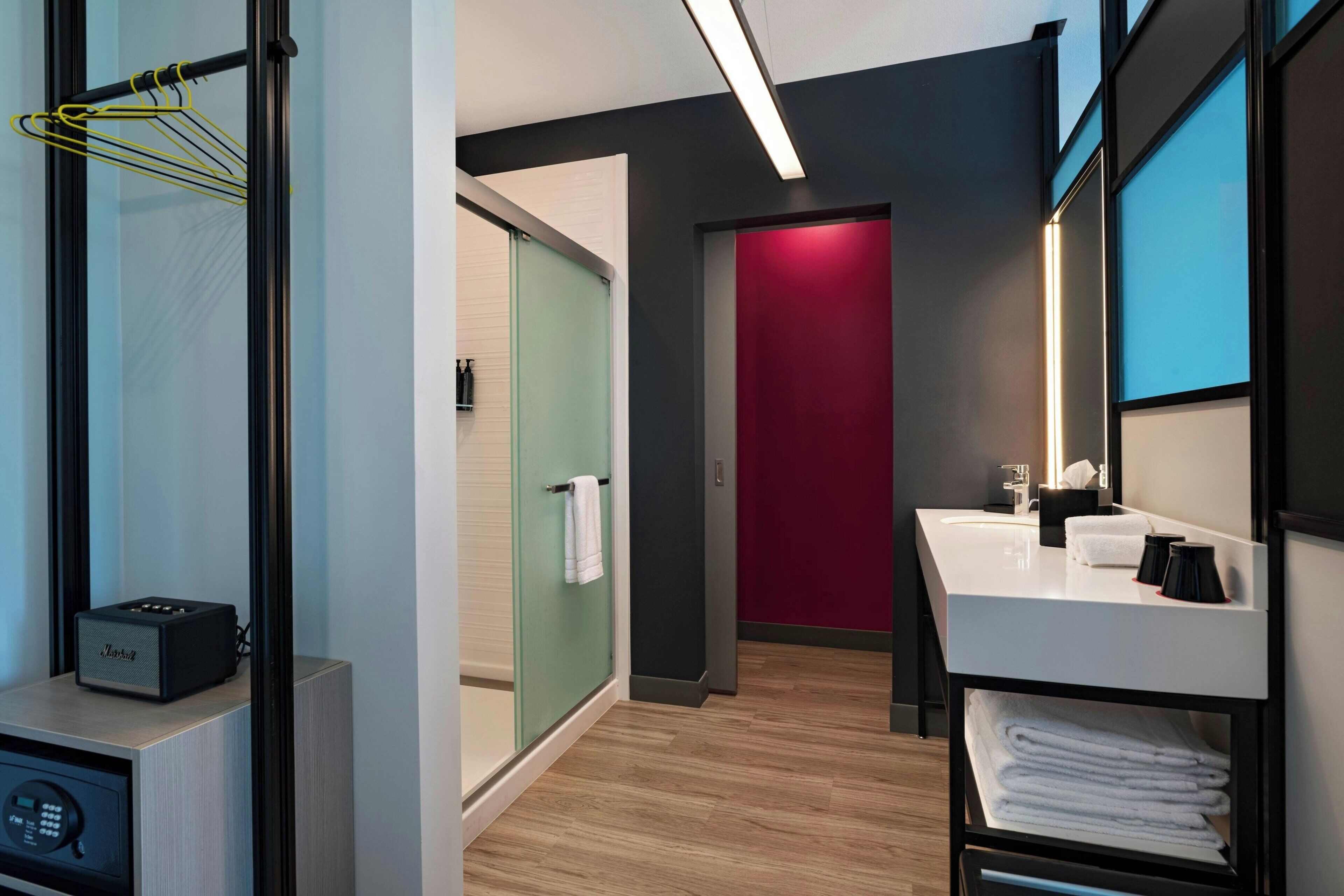 A modern hotel bathroom with a wooden floor, a white sink with black soap dispensers, a red door, and a glass shower enclosure.
