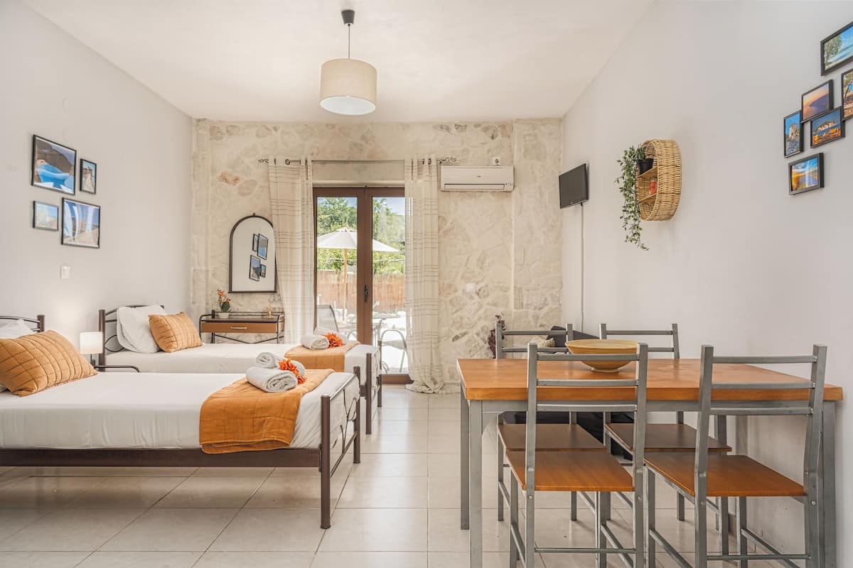 A hotel room with two beds, a dining table with chairs, a hanging light fixture, a wall-mounted TV, and a window with curtains.