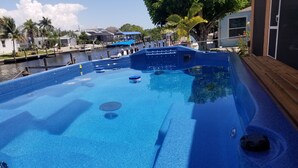 A swimming pool with a wooden deck and a view of a waterway with boats and houses in the background. - Beautiful Matlacha Isles direct access waterfront home with dock, lift and deck (Matlacha)