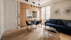 A modern living room with a dark blue sofa, a glass coffee table, and a dining area with a wooden table and chairs. -  HIGHSTAY - Luxury Serviced Apartments - Rue de Rivoli (Paris)