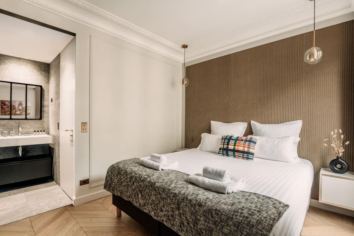 A modern hotel room with a large bed, a bedside table, a nightstand, a bathroom with a mirror, and a towel rack.