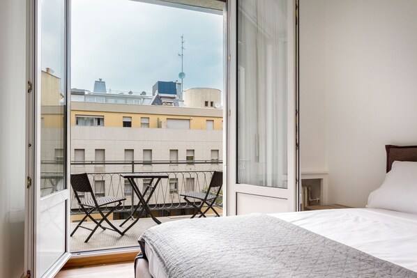 A hotel room with a bed, a small table, two chairs, and a view of the cityscape through a large window.