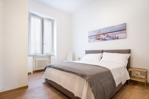 A bedroom with a bed, a window, a radiator, a bedside table, and a wall-mounted picture.
