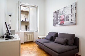 A modern living room with a grey sofa, a white desk with a computer, a lamp, and a window with a view of a balcony.