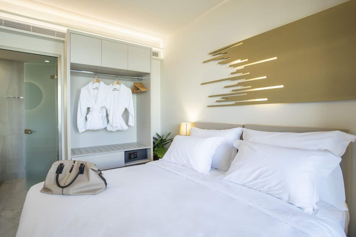A hotel room with a bed, a wardrobe, a mirror, a hanging robe, a bedside table, and a wall-mounted light fixture.